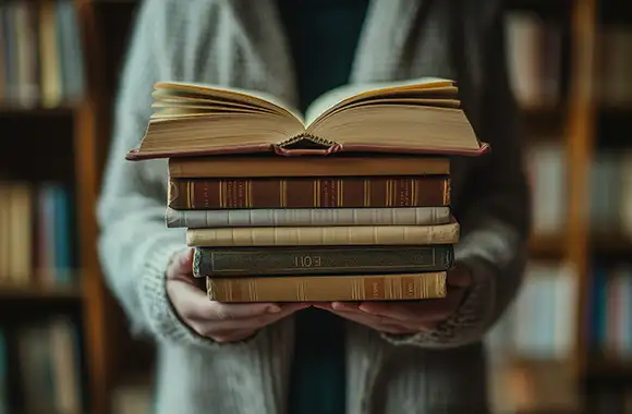 Persona sosteniendo una pila de libros relacionado a estudiar antropología social, uno de ellos abierto, en un ambiente tipo biblioteca o cafetería.