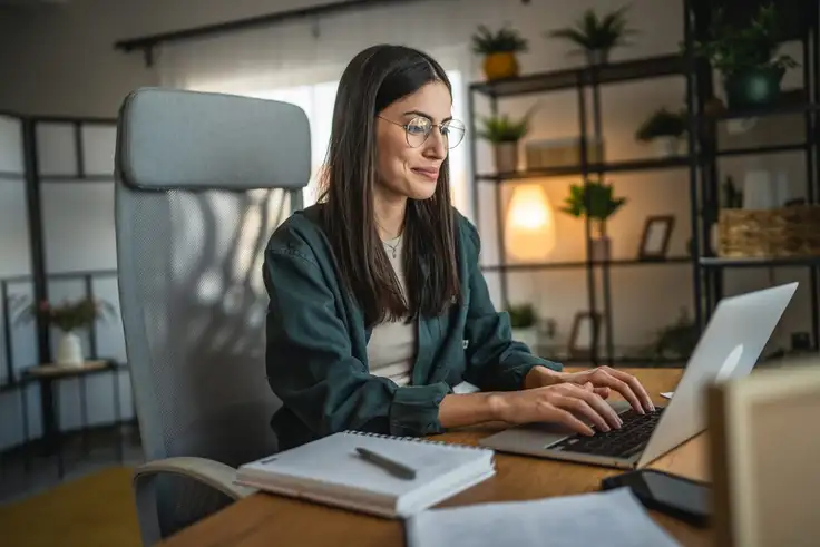 Mujer estudiando en línea desde casa con computadora portátil, rodeada de libros y tomando notas, representando las ventajas y consejos para tener éxito en la educación virtual.