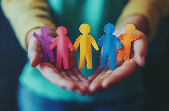 Manos sosteniendo figuras de personas de papel de colores, representando diversidad, inclusión, igualdad y la defensa de los derechos humanos, con un fondo desenfocado natural.