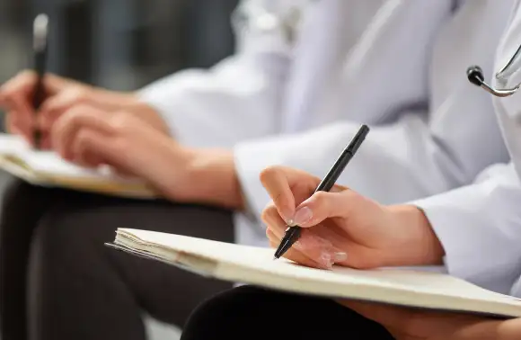 Profesionales de la salud con bata médica sentadas tomando notas en cuadernos durante una sesión de formación, relacionadas con el estudio y la práctica en salud mental.