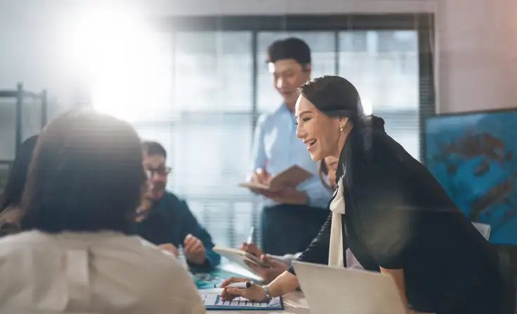 Una mujer sonriente lidera una reunión en una oficina iluminada; se inclina sobre la mesa mientras interactúa con su equipo, generando un ambiente de colaboración con liderazgo ético. 